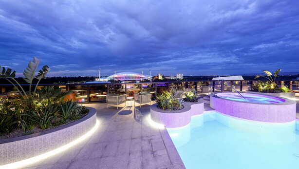 A rooftop pool area with seating and plants, a large pool, and a view of a stadium in the background. - Eos by SkyCity (Adelaide)