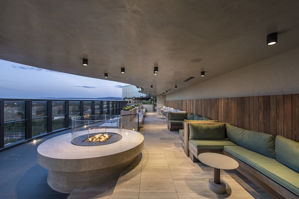 A modern rooftop lounge with a circular fire pit, green seating, and a view of the cityscape. - Eos by SkyCity (Adelaide)