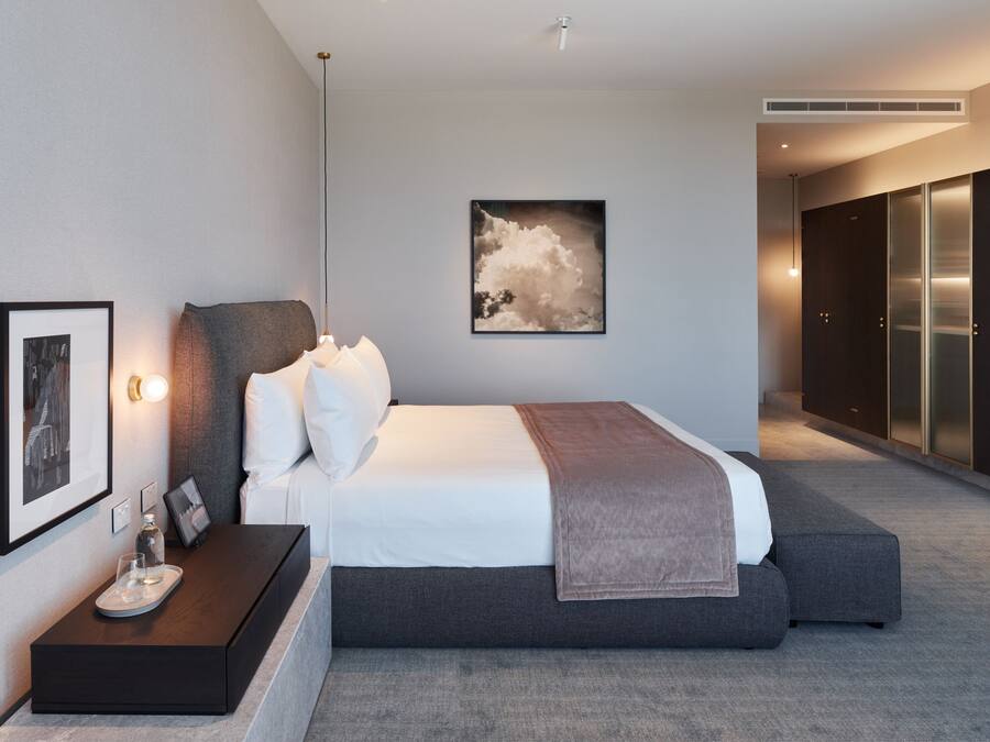 A modern hotel room with a large bed, a bedside table with a glass, and a framed picture on the wall.