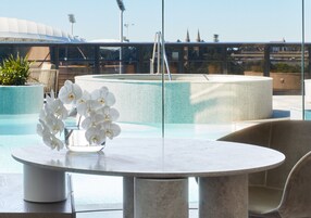 Une table ronde en marbre, ornée d’un vase rempli d’orchidées blanches, une balustrade en verre et, en arrière-plan, une vue sur un stade et des immeubles.