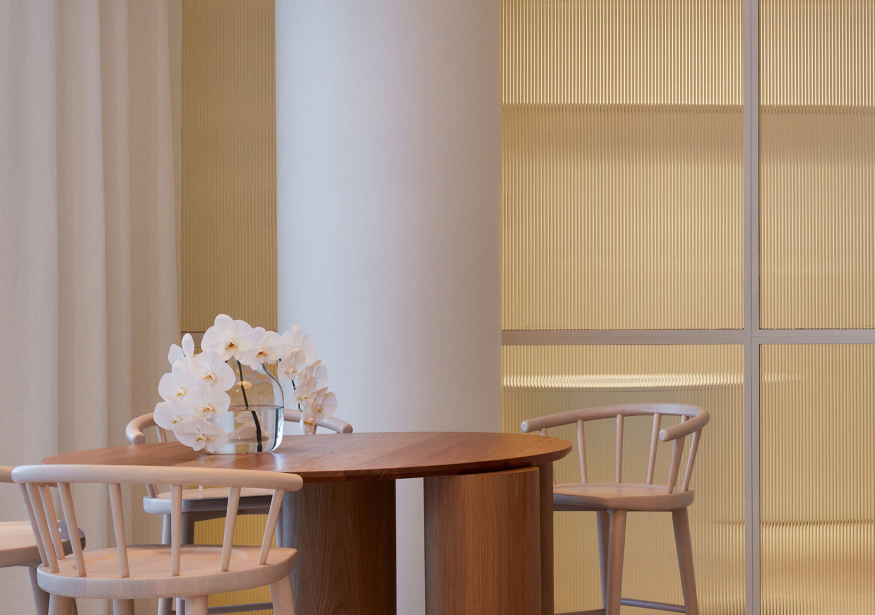 A round wooden table with two chairs, a vase with white flowers, and a textured wall in the background.