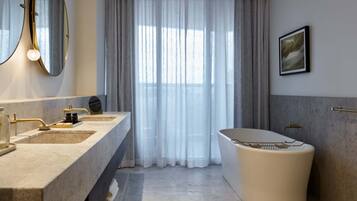 A modern bathroom with a large bathtub, two sinks with gold fixtures, a large mirror, and a window with sheer curtains.