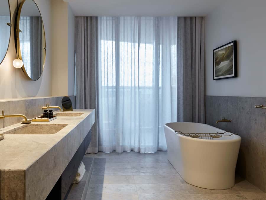 A modern bathroom with a large bathtub, two sinks with gold fixtures, a large mirror, and a window with sheer curtains.