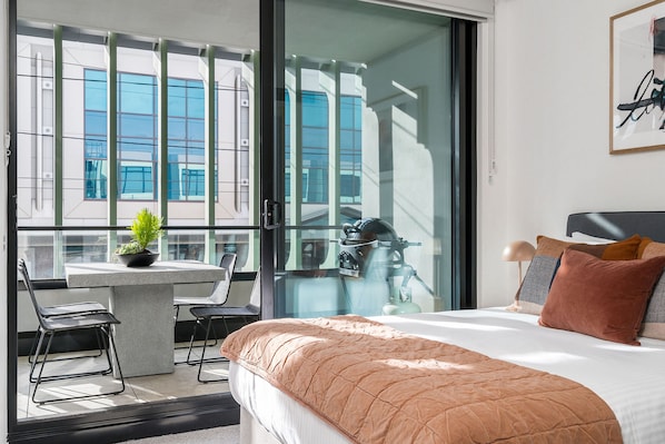 A hotel room with a bed, a small table with a plant, a chair, and a view of the city through large windows.