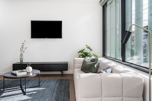 A modern living room with a flat-screen TV mounted on the wall, a black console table, a round coffee table, a beige sofa with cushions, a potted plant, and a large window with a view of the city.