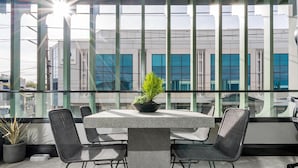 A modern outdoor seating area with a table and chairs, a potted plant, and large windows offering a view of the cityscape. - Axel Apartments 101 The Anderson (Glen Iris)