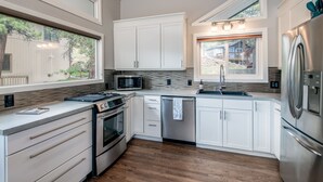 A modern kitchen with white cabinets, stainless steel appliances, a wood floor, and large windows offering views of the outdoors. - Rocky Retreat: Relax in hot tub, 2.7 mi to RMNP, walking distance to town, #3370 (Estes Park)