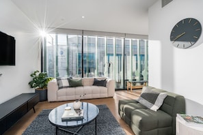 A modern living room with a sofa, a green armchair, a coffee table, and a large window with a view of buildings. - Axel Apartments 201 The Clarke (Glen Iris)