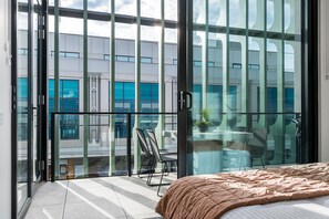 Een moderne hotelkamer met een grote schuifpui die leidt naar een balkon, een bed met een bruine dekbedovertrek en een klein tafeltje met stoelen.