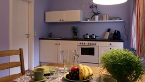 Een keuken met witte kastjes, een fornuis en een aanrecht. Er staat een tafel met een karaf, een fruitschaal en een groene plant.