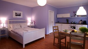 Een slaapkamer met een bed, nachtkastjes, een stoel, een eettafel met stoelen, een keuken met kastjes, een fornuis en een hanglamp.