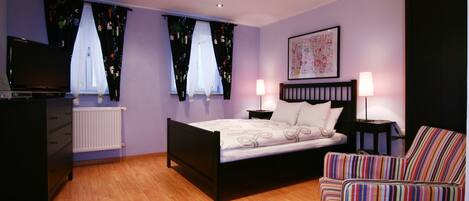 Une chambre à coucher avec un lit, deux tables de chevet avec des lampes, une télévision, une commode et un fauteuil à rayures.