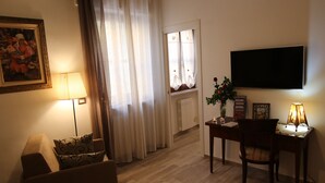 A living room with a sofa, a table with a lamp, a TV mounted on the wall, and a window with curtains. - DOMUS LEVI APARTMENT (Matera)