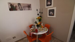 A dining area with a round table, orange chairs, and a variety of food and drink items. There are wall decorations and a framed picture above the table. - DOMUS LEVI APARTMENT (Matera)