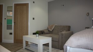 A room with a wooden door, a white table with a plant, a brown sofa, and a bed with a white headboard. - Frinton Escapes (Frinton-on-Sea)