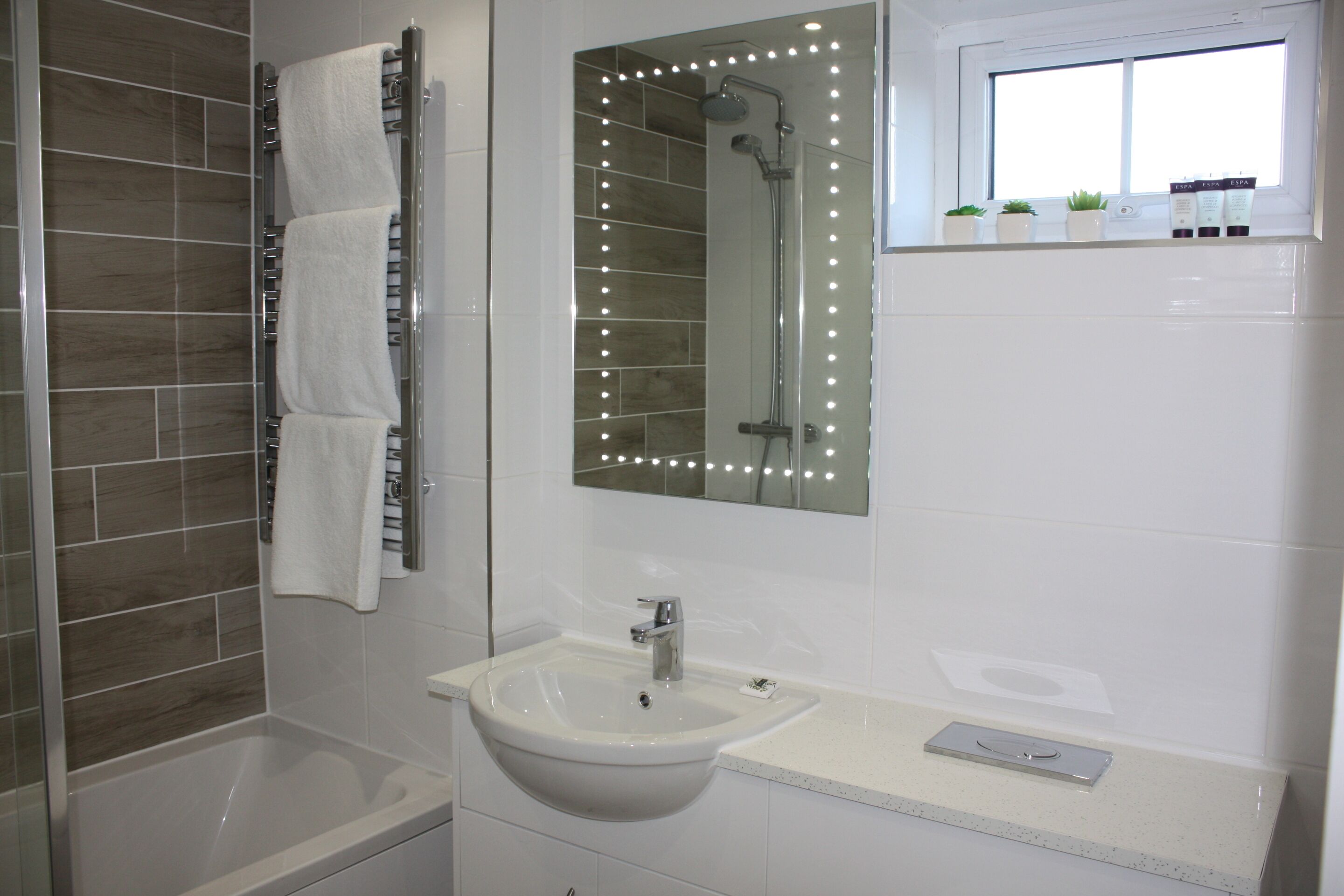 Un baño con bañera, lavamanos, espejo con iluminación y ventana con plantas.