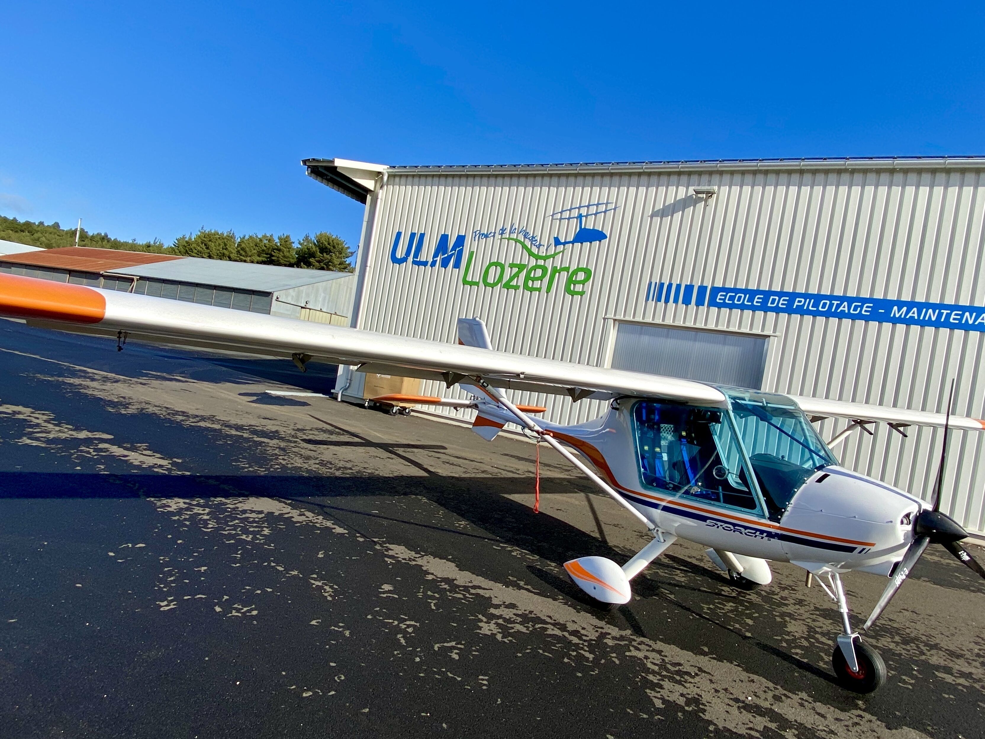 A small aircraft parked in front of a hangar with the text "Ulm Lozere" and "Ecole de Pilotage - Maintenance".