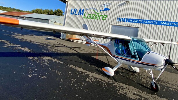 Un petit avion est garé devant un hangar sur lequel on peut lire les inscriptions « Ulm Lozère » et « École de pilotage - Maintenance ».