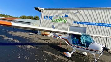 A small aircraft parked in front of a hangar with the text "Ulm Lozere" and "Ecole de Pilotage - Maintenance".