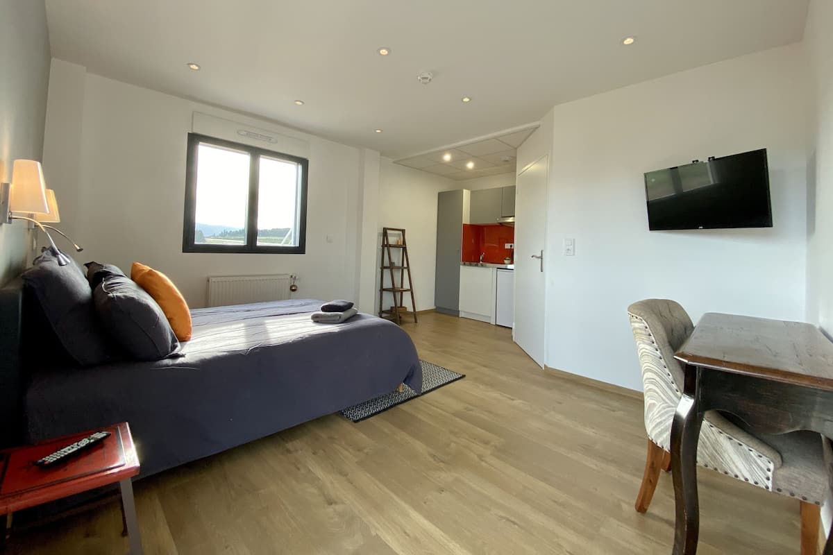 Un dormitorio con cama, escritorio, silla, televisión y vista a una cocina.