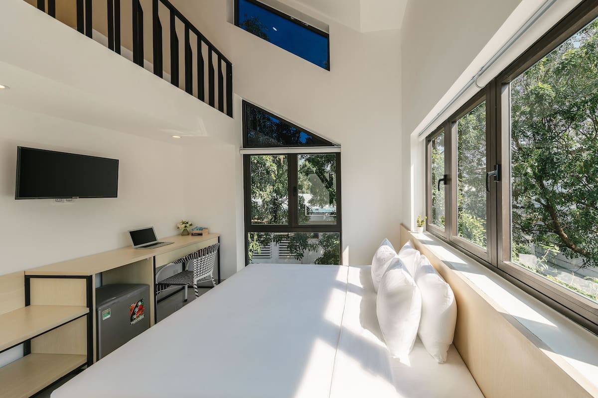 A modern hotel room with a bed, a desk, a TV, and large windows offering a view of trees.