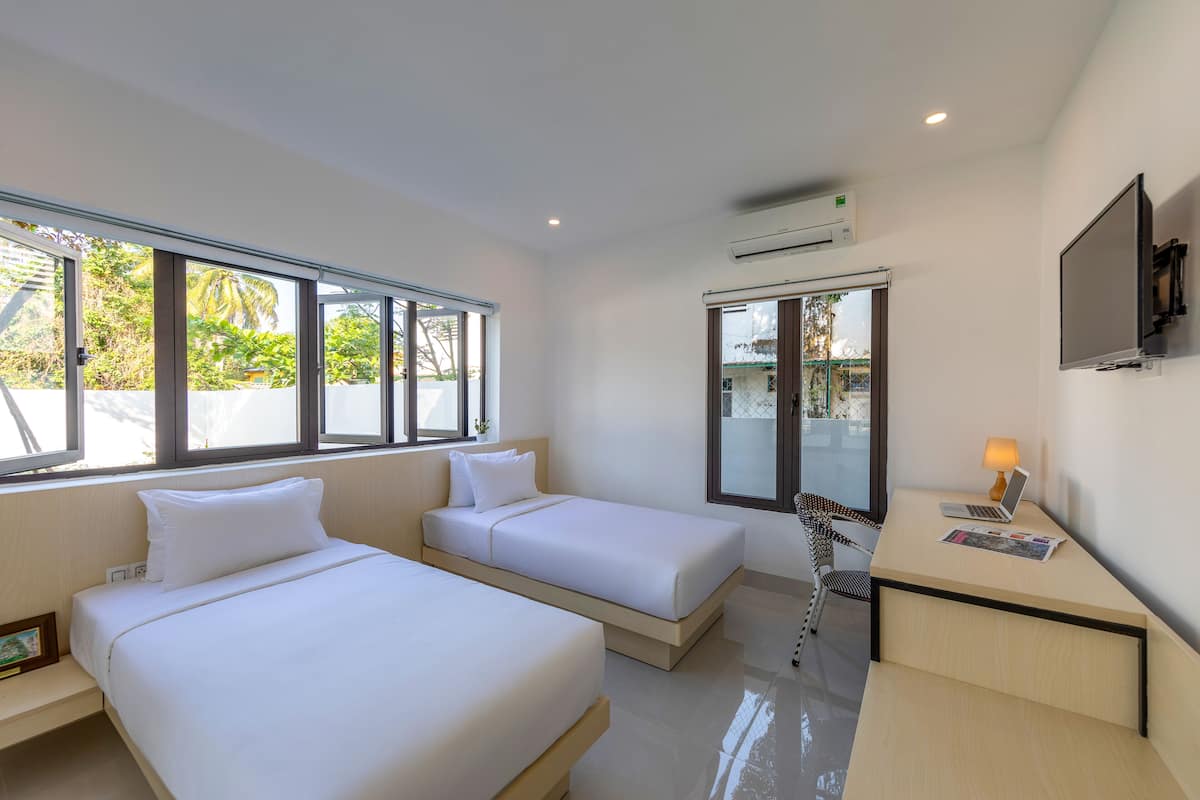 A modern hotel room with two single beds, a desk with a laptop and lamp, a TV, and large windows offering a view of greenery outside.