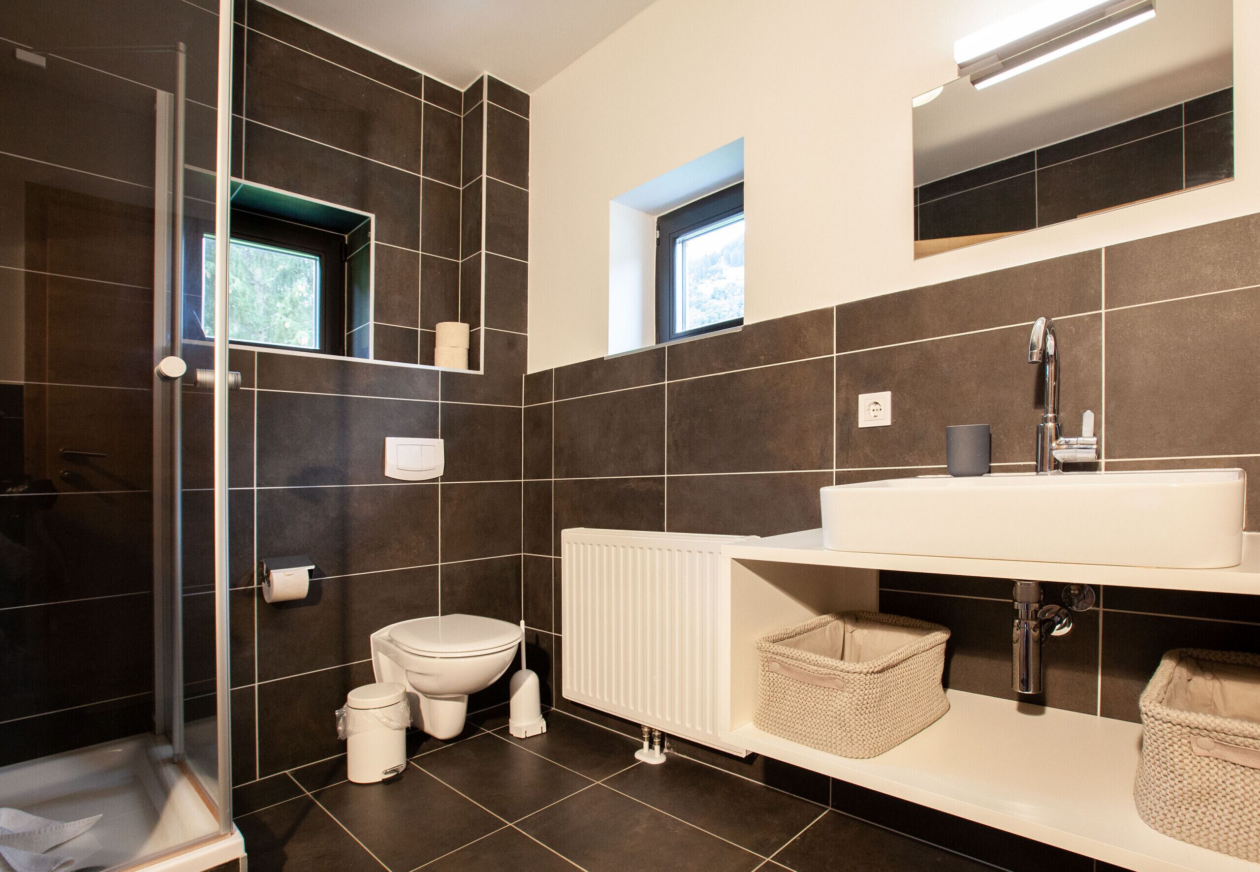 Ein modernes Badezimmer mit Duschkabine aus Glas, Toilette, Waschbecken mit Spiegel und einem Fenster für natürliches Licht.