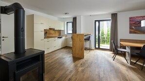 A modern kitchen with a wood-burning stove, a dining area with a wooden table and chairs, and a large window with a view of greenery. - 4513 Alpin 13 (St. Gallenkirch)