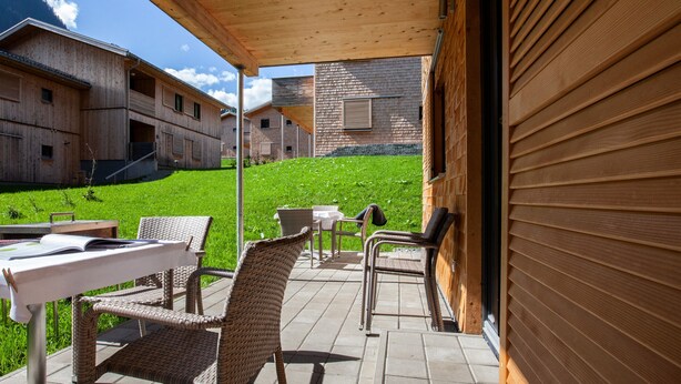 Een overdekt terras met rieten meubels en een tafel, met uitzicht op een groen gazon en houten gebouwen.
