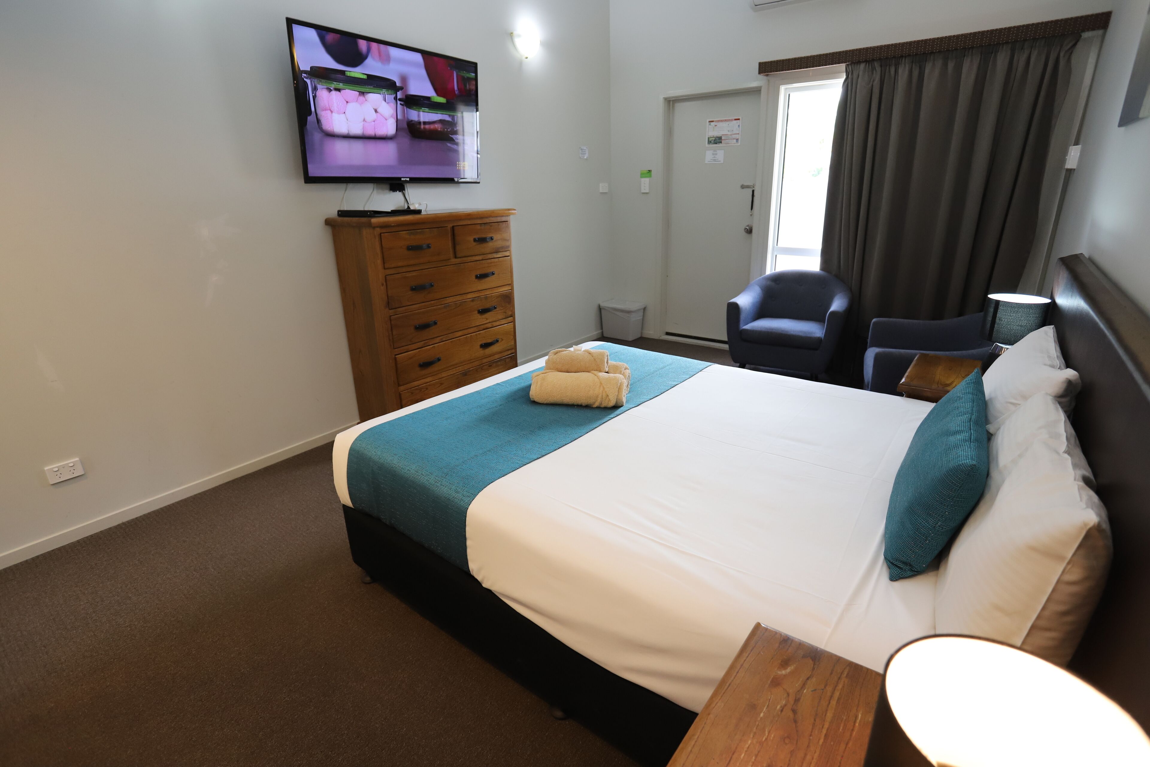 Una habitación de hotel con cama, televisor montado en la pared, cómoda de madera, silla y mesita de noche con lámpara.