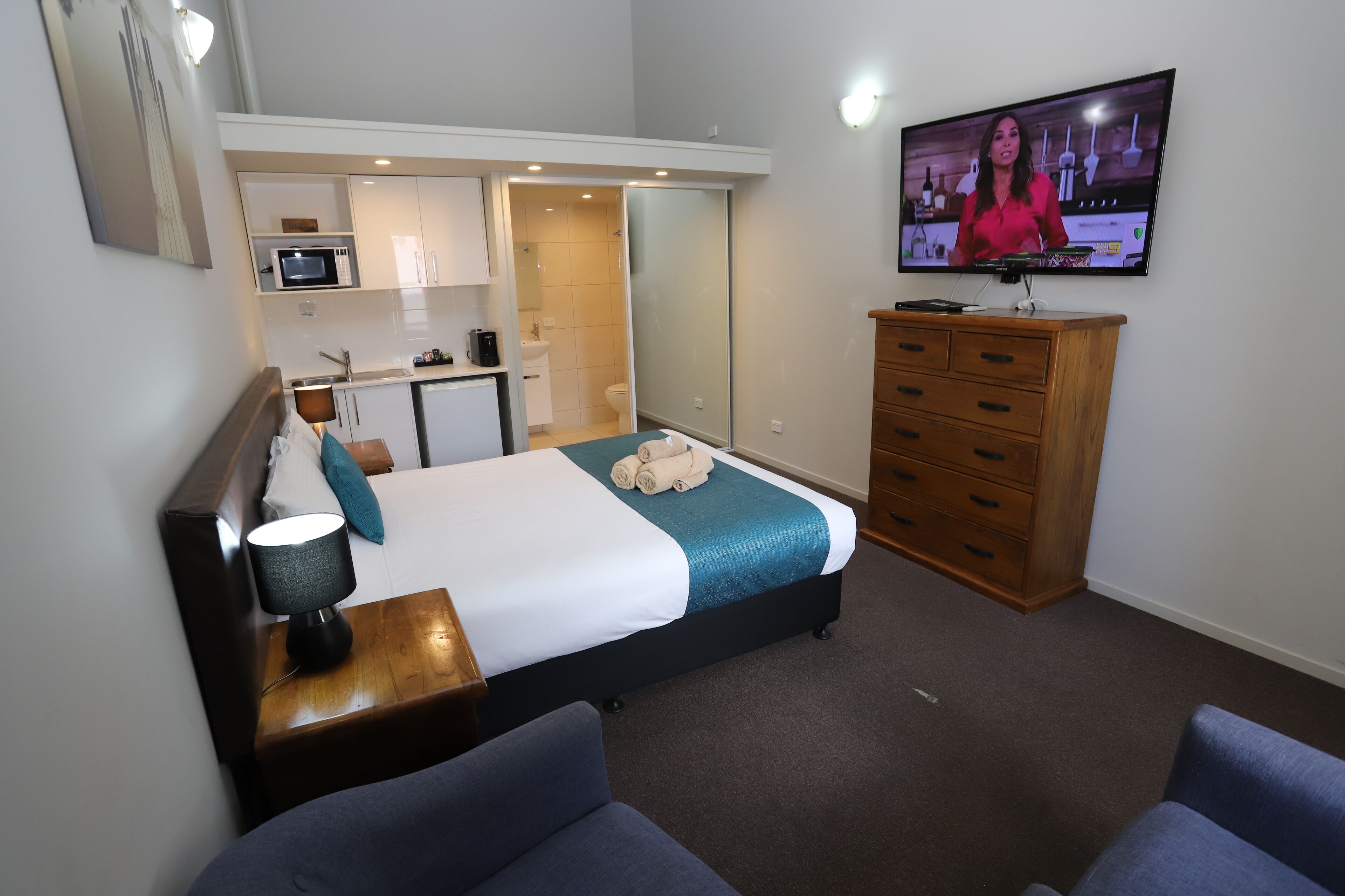 A hotel room with a bed, a TV, a desk, a chair, a dresser, and a kitchenette.