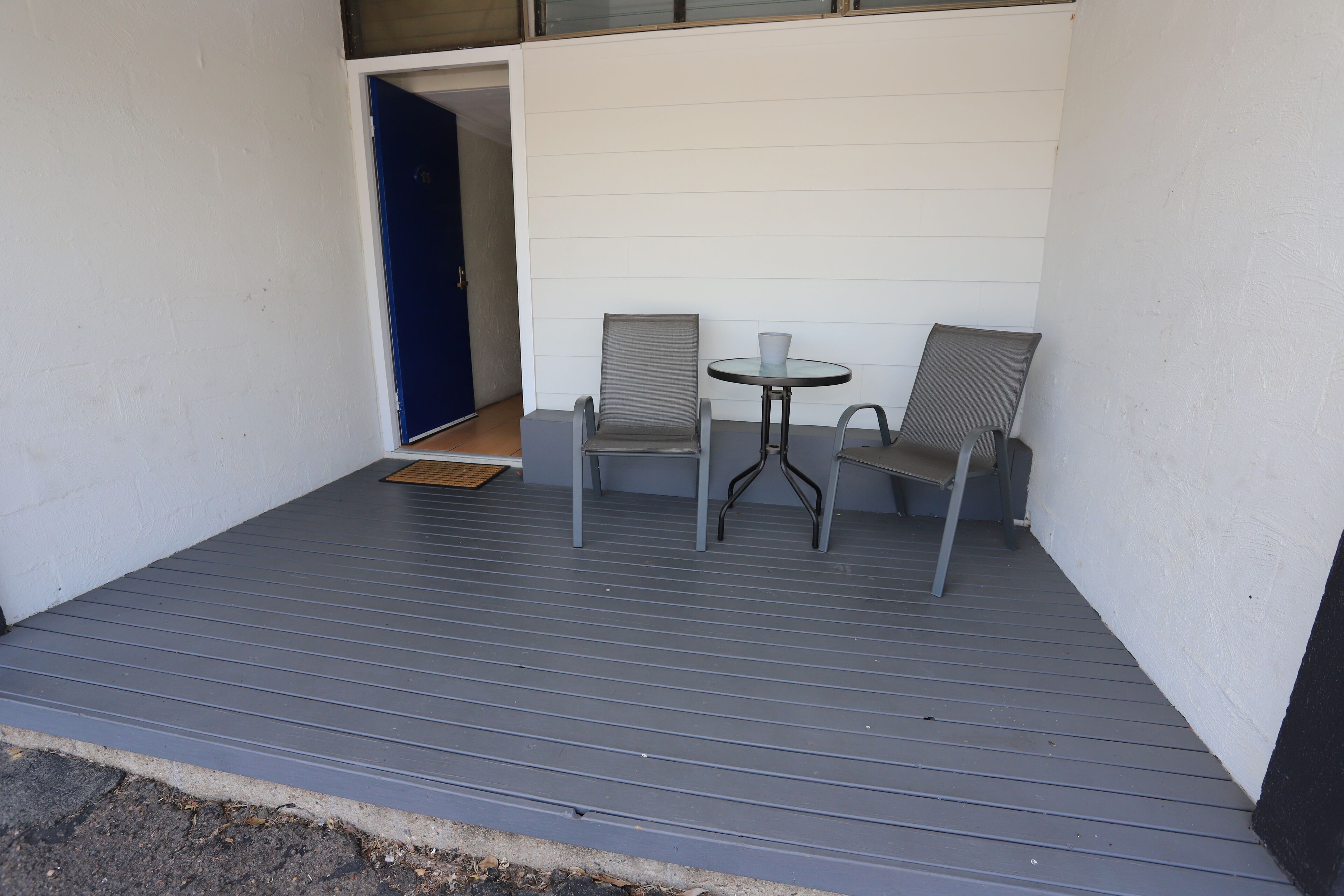 A covered outdoor seating area with two chairs and a table, a door, and a white wall.