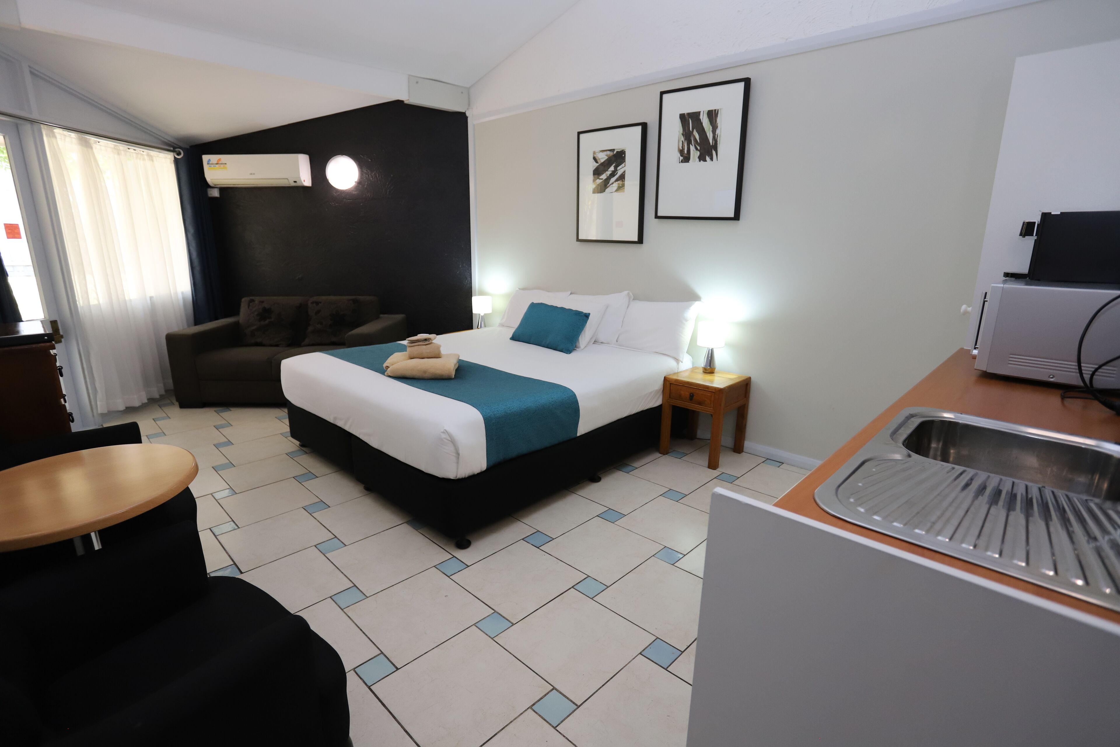 Een hotelkamer met een bed, nachtkastje, lamp en een kitchenette.