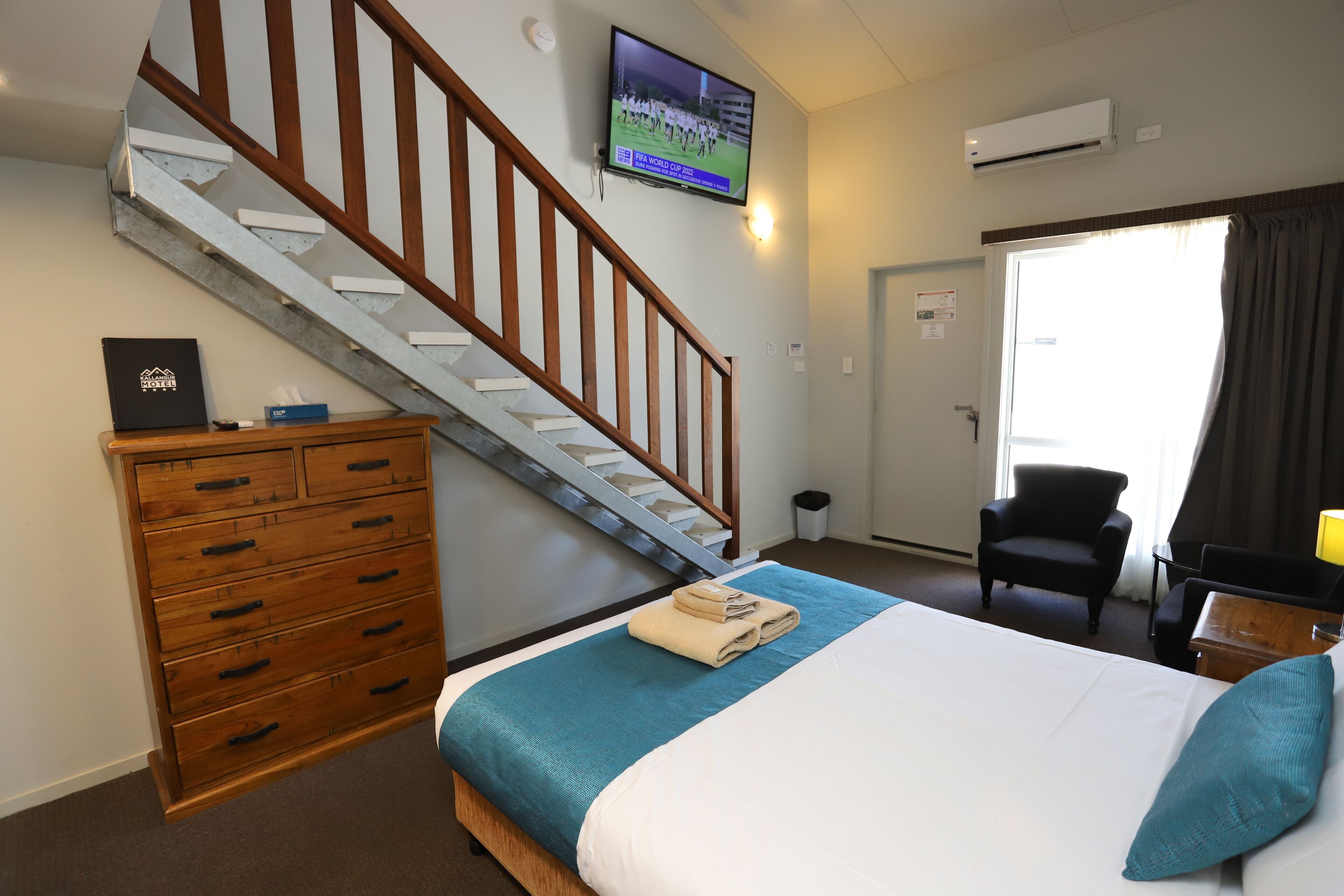 Camera d'albergo con un letto, una cassettiera in legno, un televisore a parete, una scala e una porta che dà all'esterno.