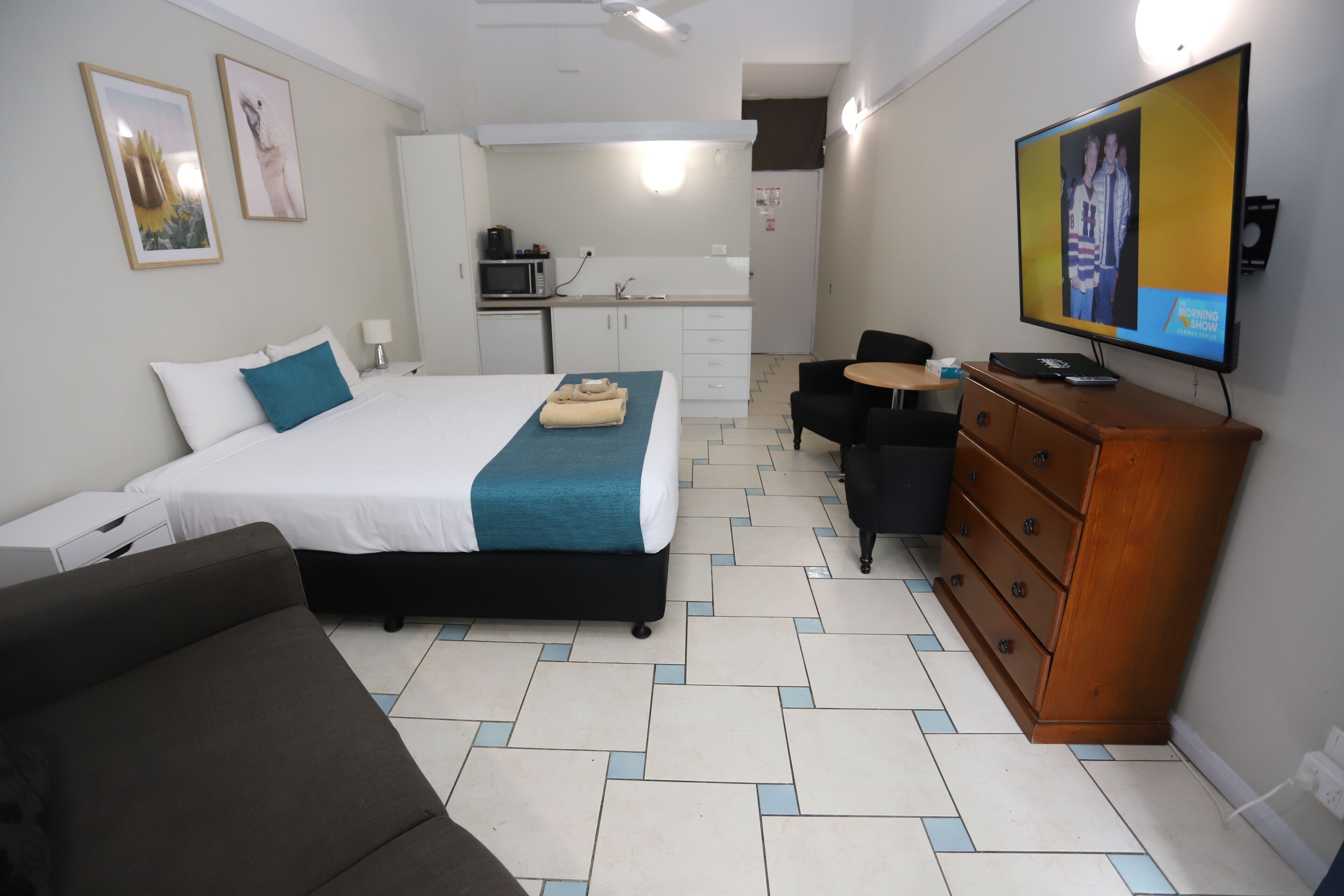 A hotel room with a bed, a sofa, a TV, a dresser, and a kitchenette in the background.