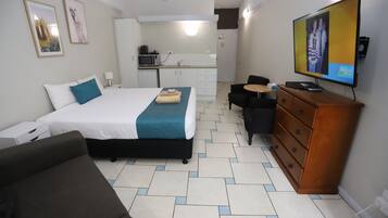 A hotel room with a bed, a sofa, a TV, a dresser, and a kitchenette in the background.