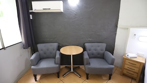 Een kamer met twee grijze fauteuils, een kleine ronde tafel, een wandairconditioner en een houten nachtkastje met lades.