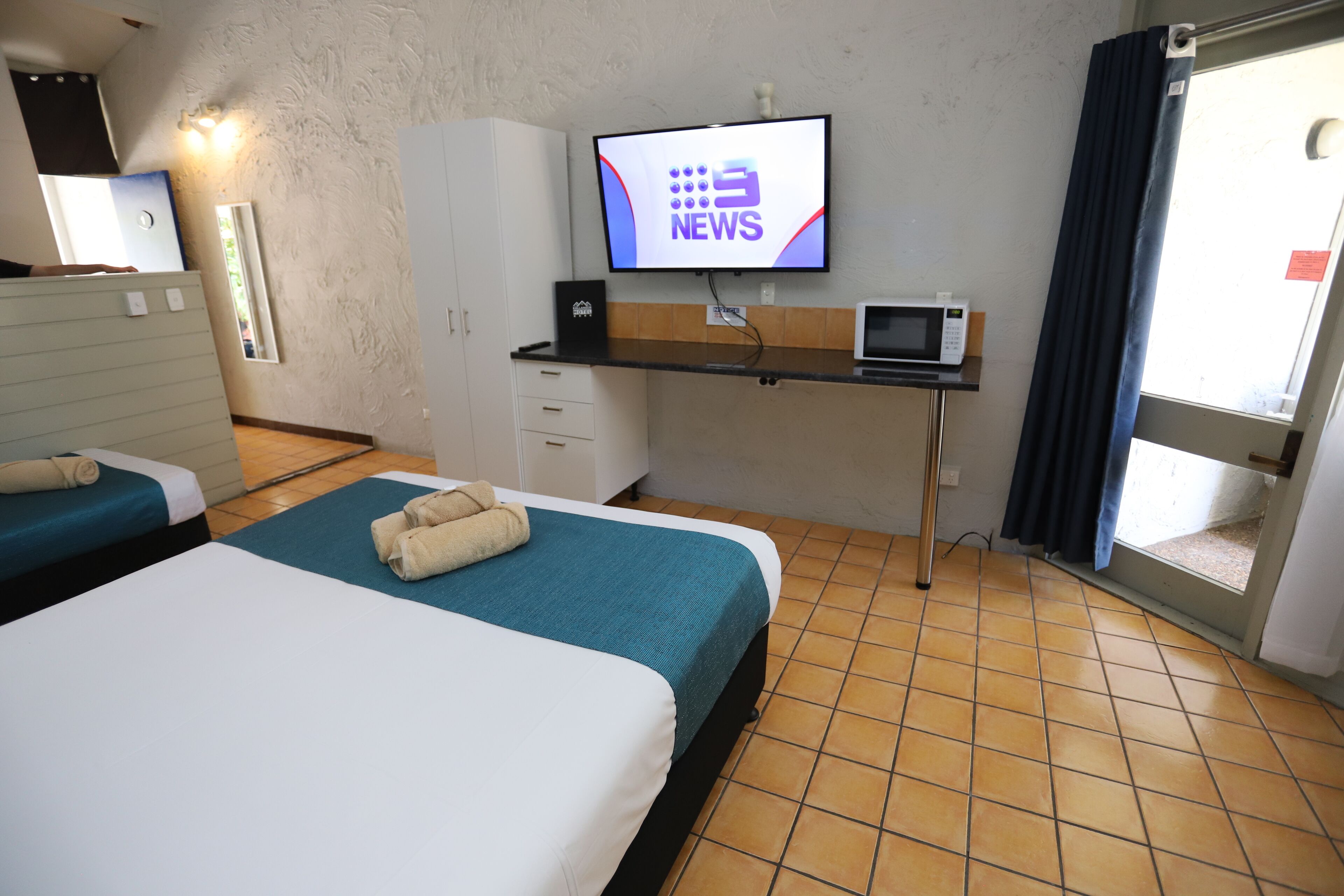 Habitación de hotel con dos camas, un escritorio con televisor mostrando un noticiero, microondas y ventana con cortinas.