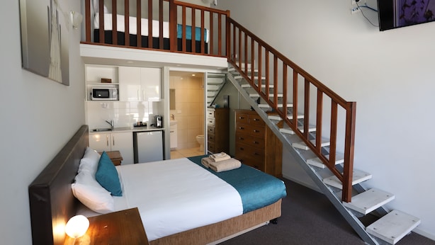 Een slaapkamer met een bed, nachtkastje, houten hoofdbord, trap, keukenhoek en badkamer.