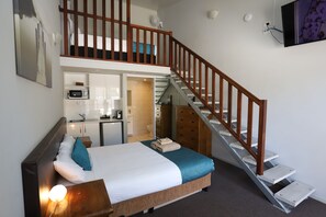 A bedroom with a bed, a nightstand, a wooden headboard, a staircase, a kitchen area, and a bathroom.