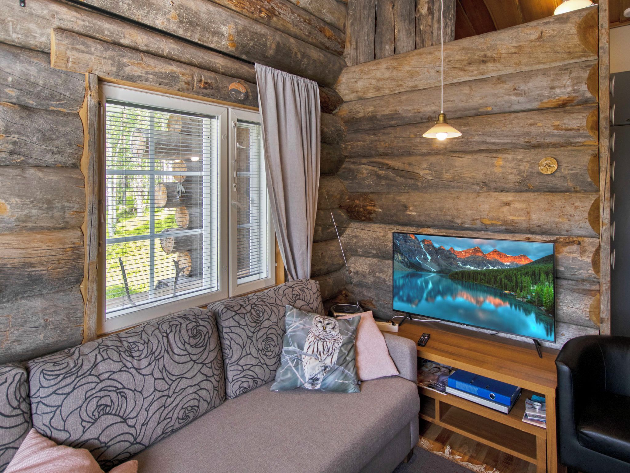 L’intérieur d’une cabane en rondins, avec un canapé, un téléviseur et une fenêtre équipée de stores.
