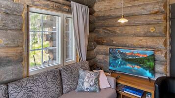 L’intérieur d’une cabane en rondins, avec un canapé, un téléviseur et une fenêtre équipée de stores.
