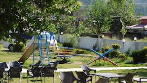An outdoor seating area with tables and chairs, a playground with slides and swings, and a view of a hilly landscape in the background. - Pine View hotel and Restaurant (Balakot)