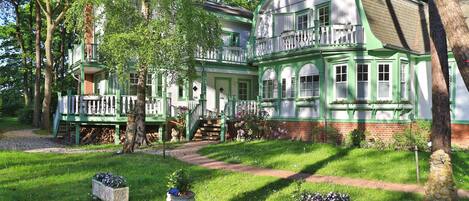 A large, two-story house with a green and white exterior, a balcony, and a well-maintained garden with flower beds.