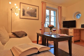 A cozy room with a sofa, a table with a book and glasses, a painting, a window with curtains, a TV, and a small round window. - Landhaus Victoria, Seestern (Ostseebad Boltenhagen)