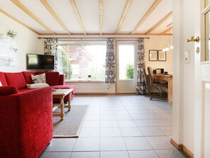 A living room with a red sofa, a TV, a dining area with a table and chairs, and a large window with curtains. - Haus Seepferdchen Whg. SP2 (Ostseebad Boltenhagen)