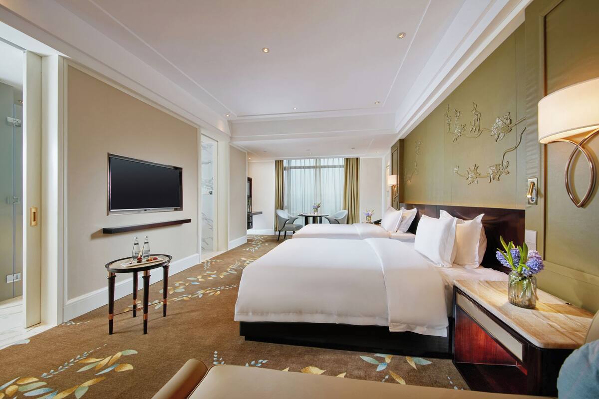 Ein Hotelzimmer mit einem großen Bett, einem an der Wand montierten Fernseher, einem kleinen Tisch mit Blumenvase und Blick ins Freie durch das Fenster.
