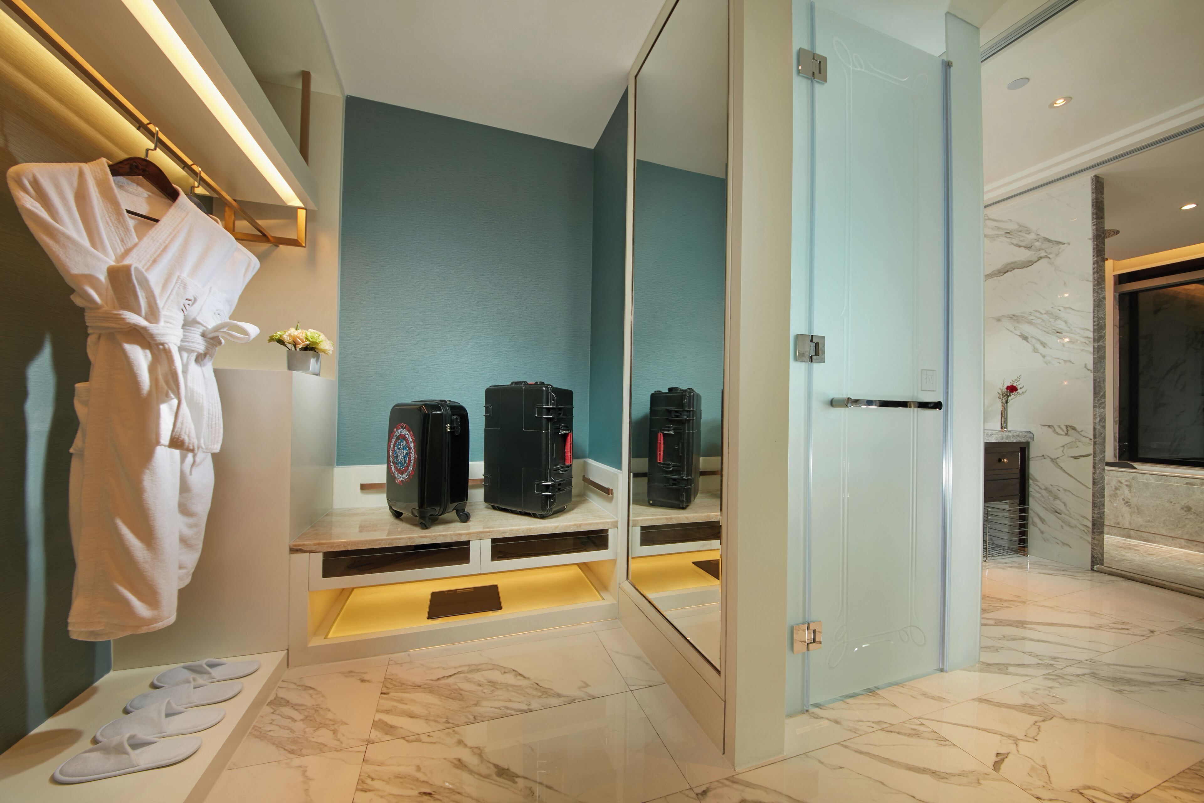 A modern bathroom with a robe hanging on a rack, a mirror, and a bench with shoes. There are two black suitcases on the bench.