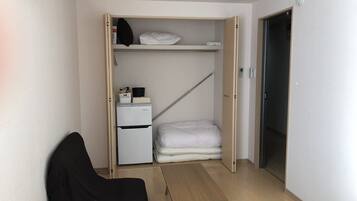 A compact room with a bed, a small desk, a wardrobe with a mirror, and a mini-fridge.