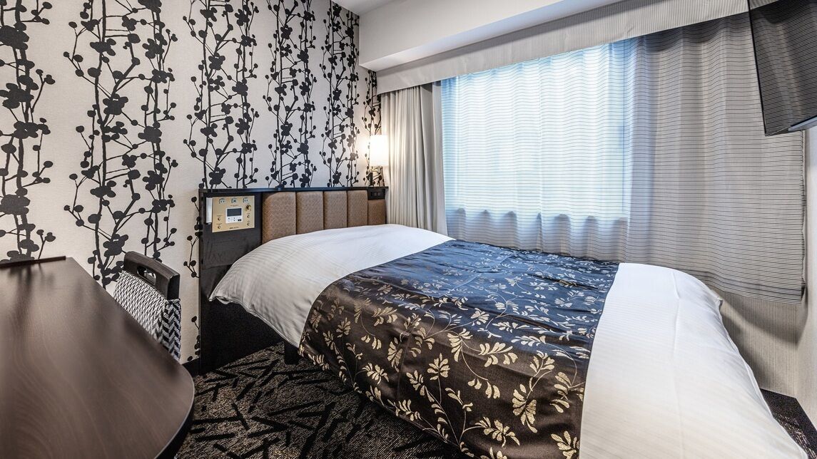 A hotel room with a bed, a desk, a nightstand, and a window with curtains.